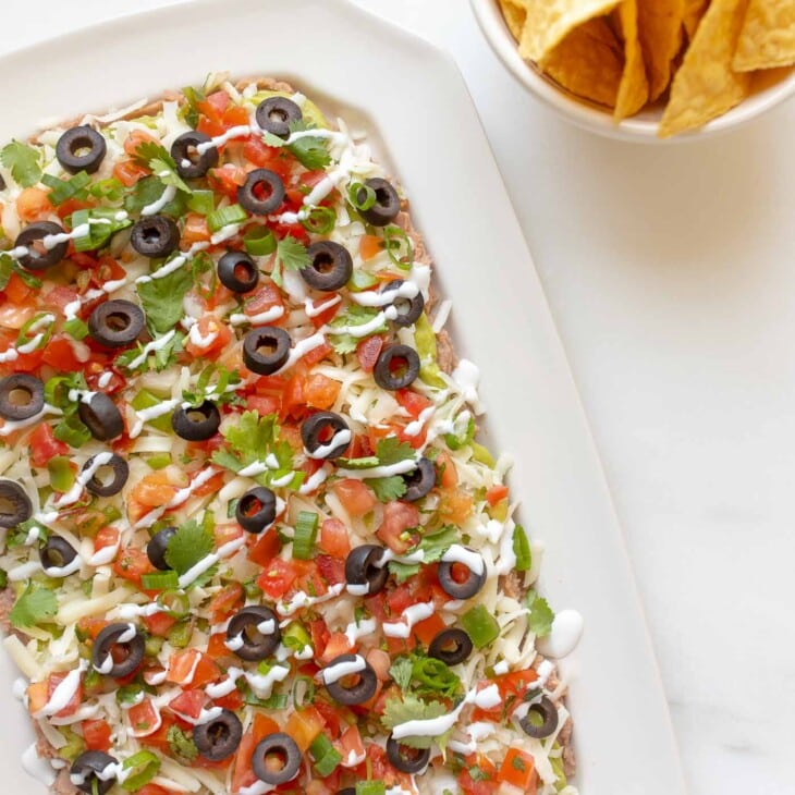 7 layer dip in a white dish, tortilla chips in a white bowl to the side.