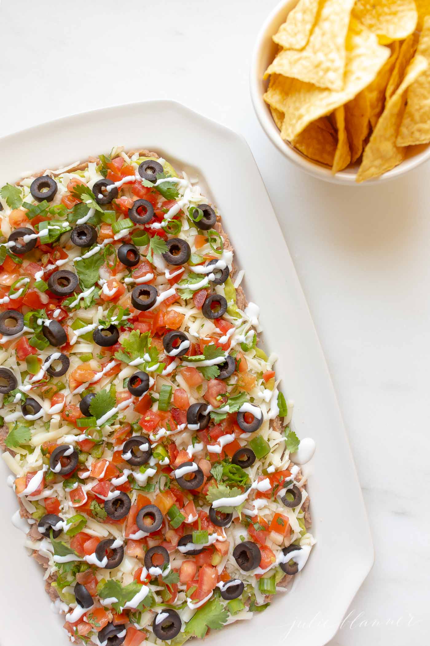 7 layer dip in a white dish, tortilla chips in a white bowl to the side.