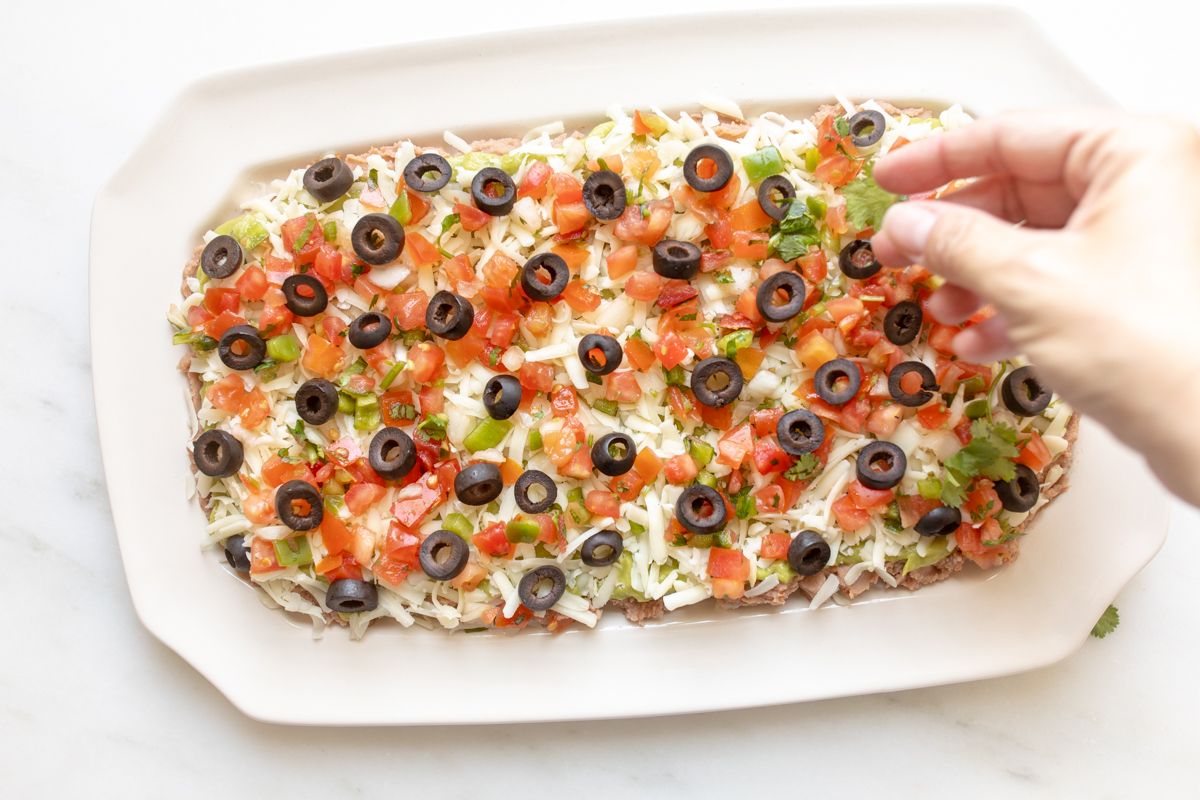 A hand adding garnish to a white platter of 7 layer dip.