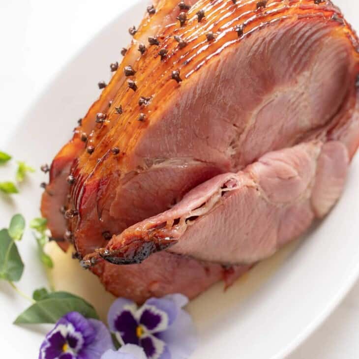 A honey ham studded with cloves on a white platter, garnished with fresh flowers for Easter