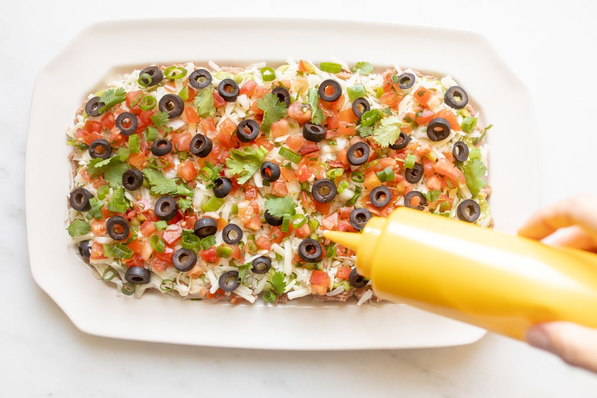 A hand adding garnish to a white platter of 7 layer dip.