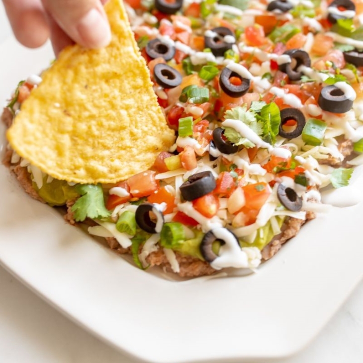 A hand dipping a tortilla chip into a platter of 7 layer dip.