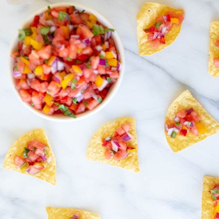 A marble countertop with tortilla chips scattered across, topped with fresh watermelon salsa. Bowl of watermelon salsa in the corner.