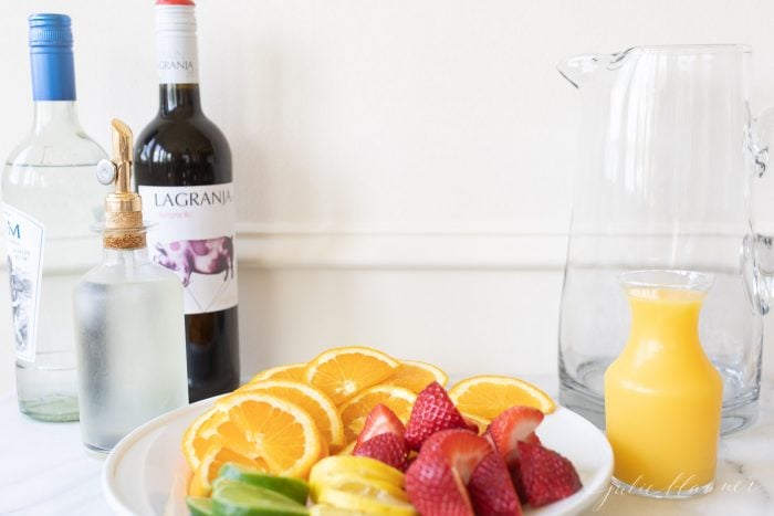 A bottle of wine with a glass pitcher and plate of various fruits on a marble surface. 