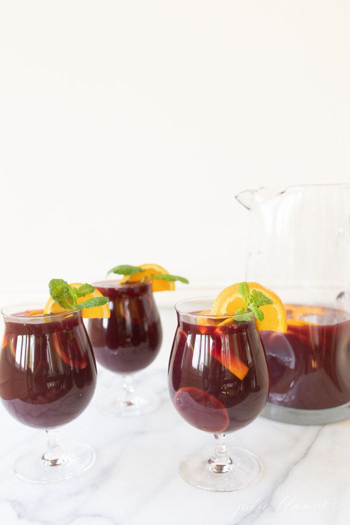 Sangria Punch in three glasses, garnished with an orange. A glass pitcher with the mixture is to the side.