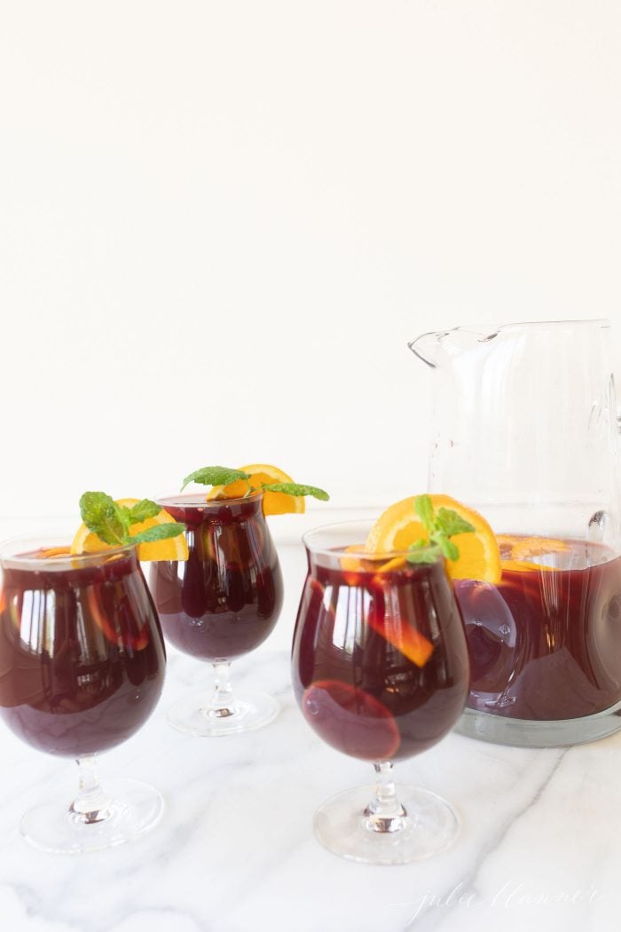 Three cups of sangria, garnished with a lemon slice and mint. A glass pitcher of sangria is to the side. 