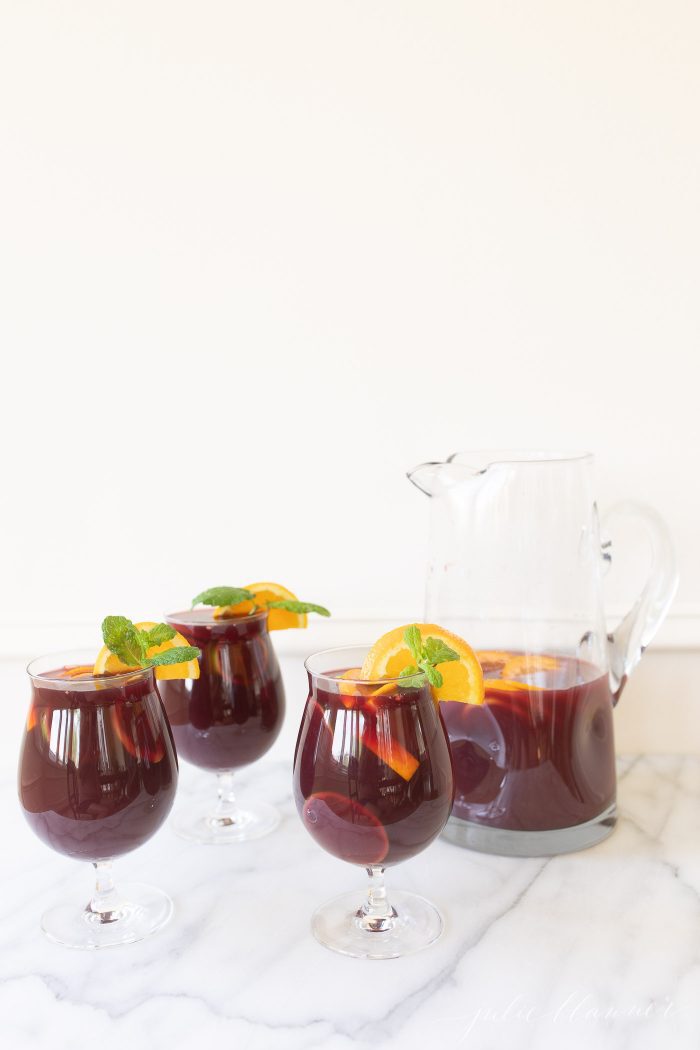 Three cups of sangria, garnished with a lemon slice and mint. A glass pitcher of sangria is to the side. 