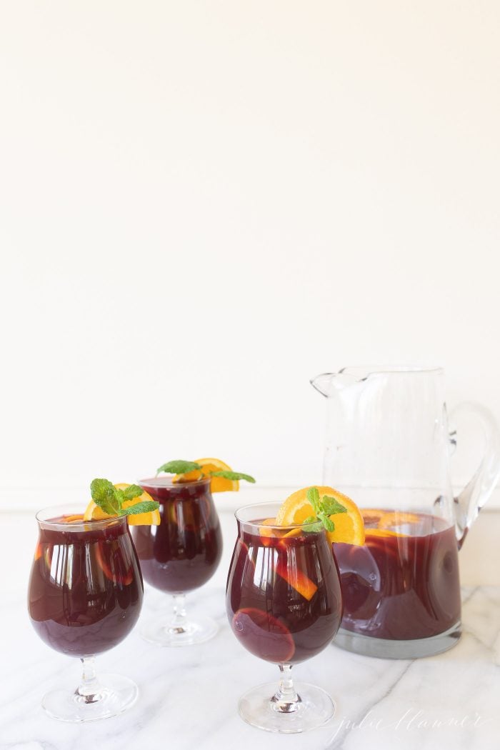Sangria Punch in three glasses, garnished with an orange. A glass pitcher with the mixture is to the side.