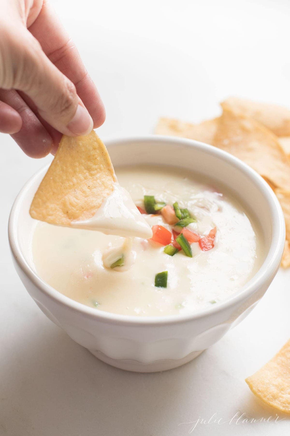 A hand reaching into a bowl of white queso with a tortilla chip, topped with pico de gallo