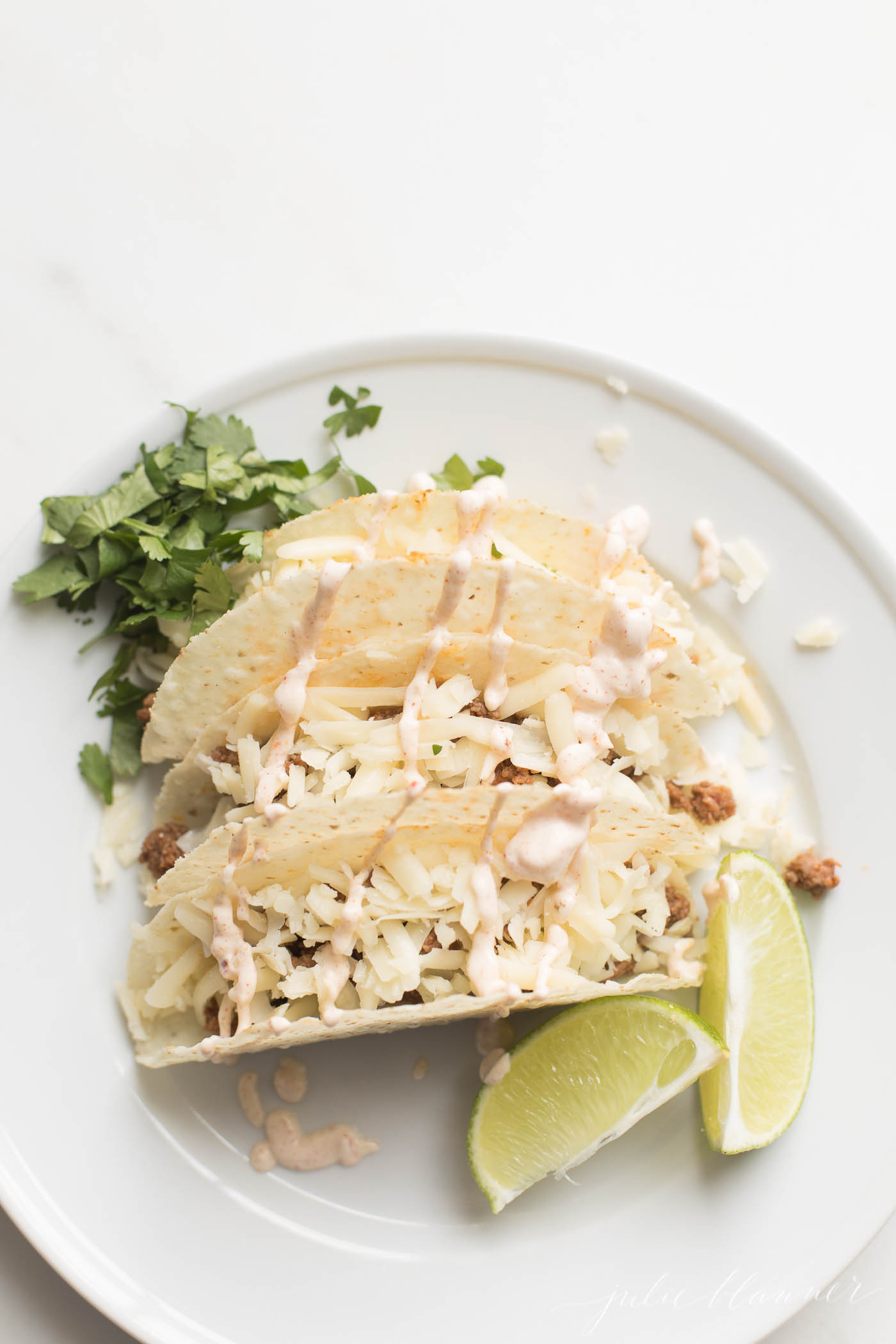 A white plate with three hard shell beef tacos topped with cheese.