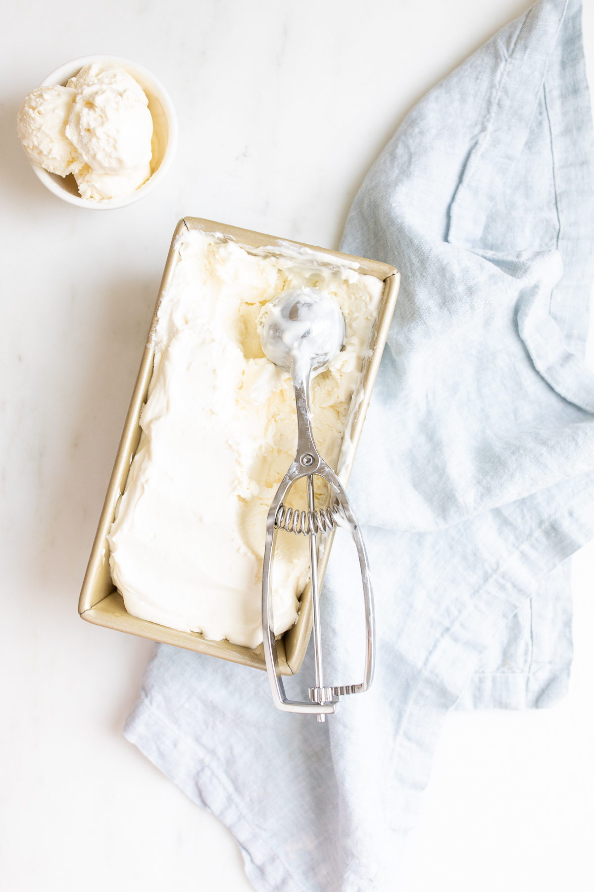 cream cheese ice cream in a loaf pan with an ice cream scooper and small white bowl of ice cream