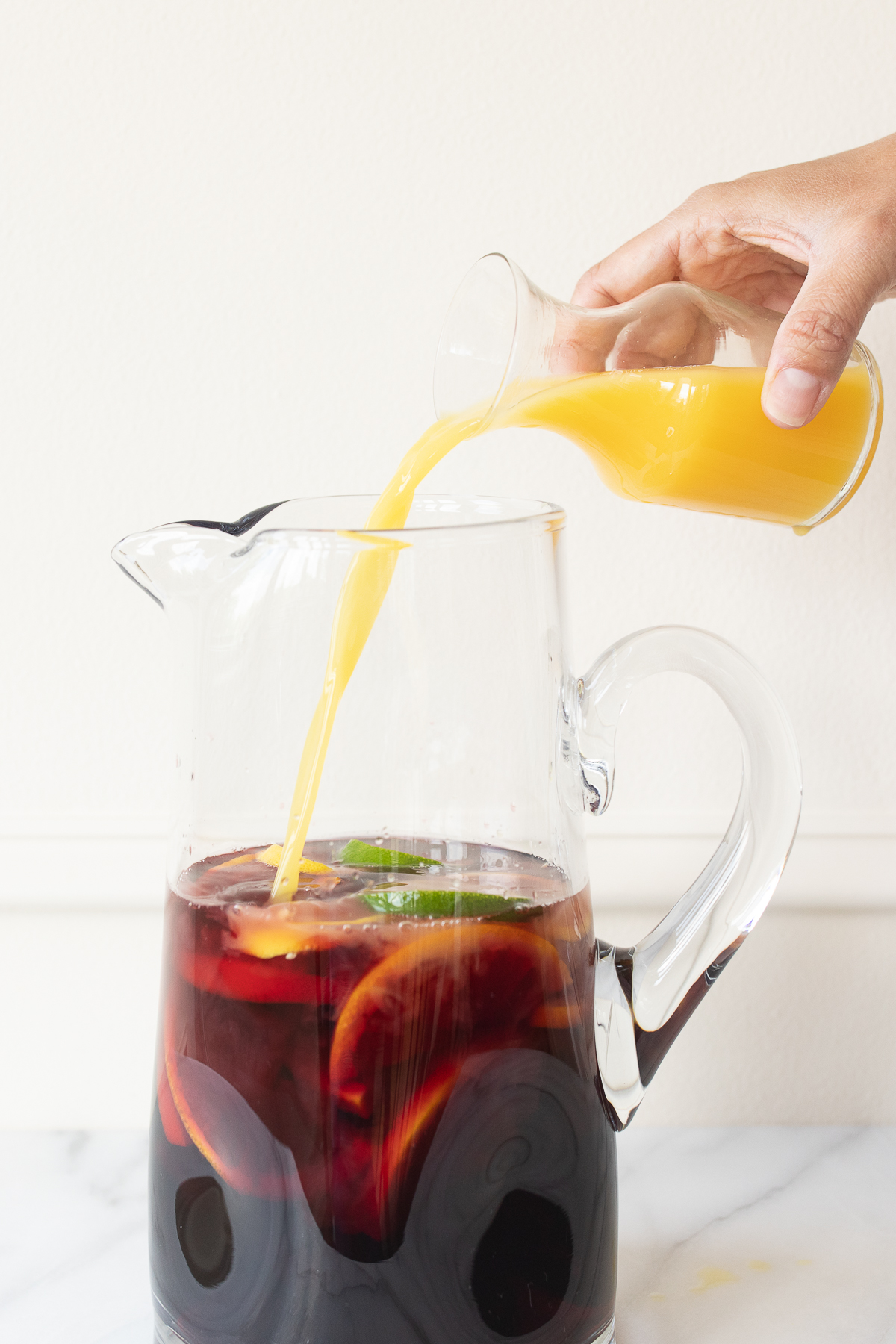 A glass pitcher full of red sangria, with orange juice being poured into it.