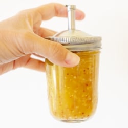 A hand holding a glass jar with a spout lid, full of pasta salad dressing