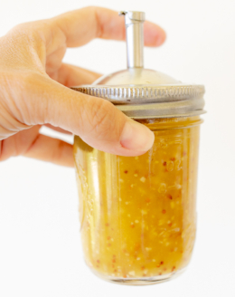 A hand holding a glass jar with a spout lid, full of pasta salad dressing