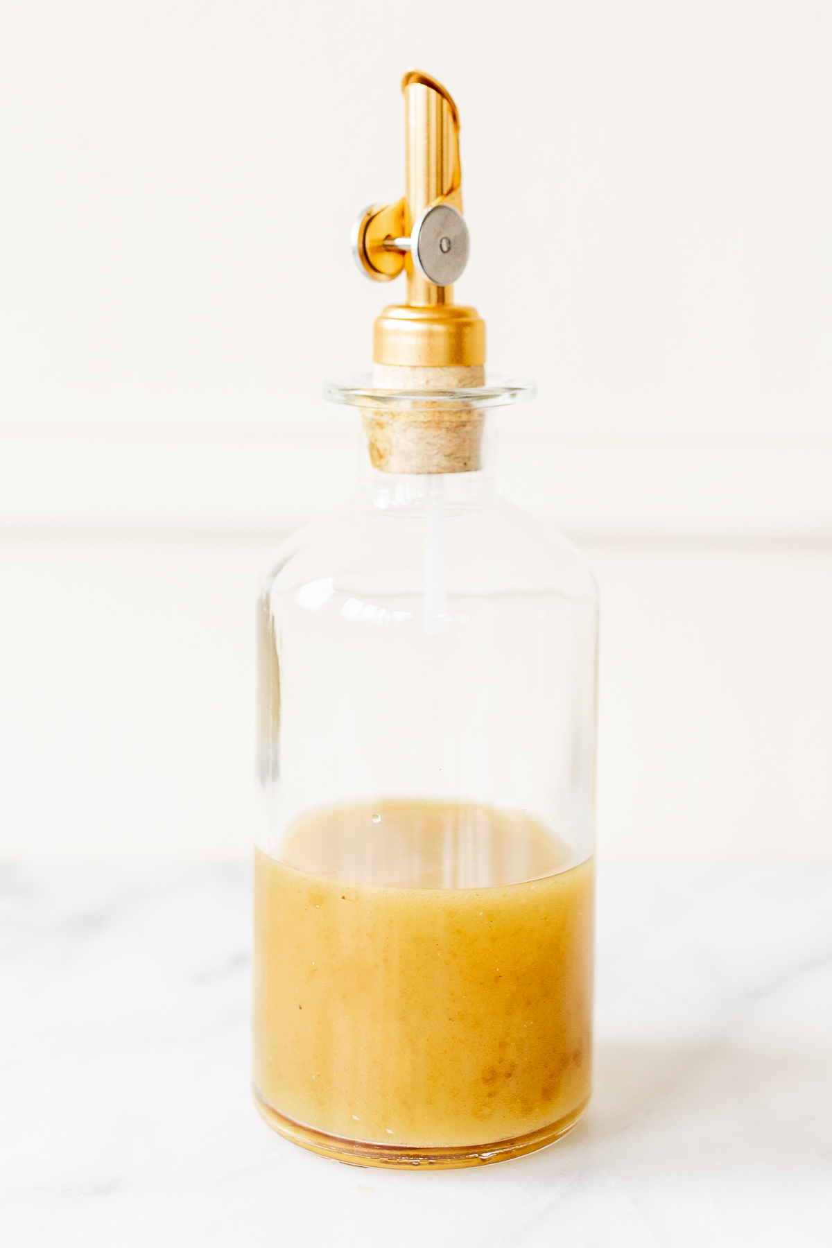 A glass bottle with a gold stopper on a marble countertop, filled with sherry vinaigrette.