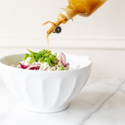 A bottle of dressing being poured over a salad in a white bowl.