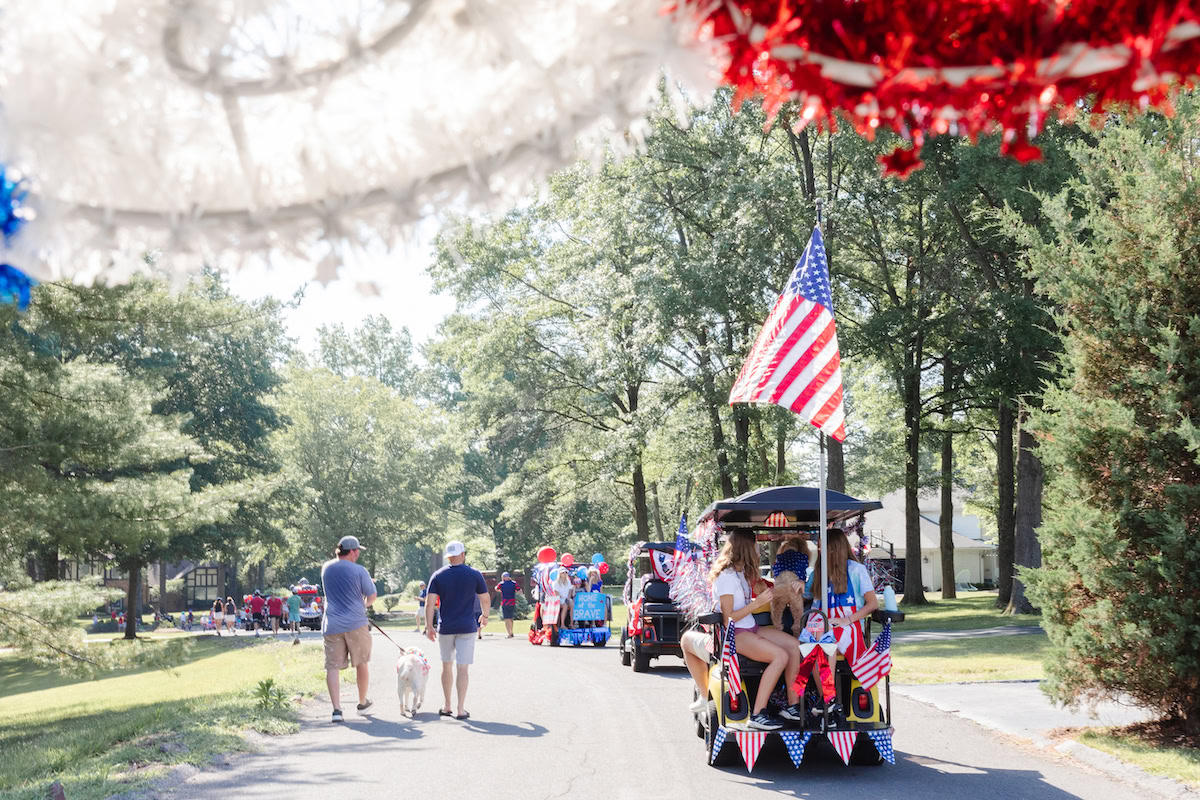 People walking and riding decorated golf carts with American flags in a tree-lined street during a daytime parade. 4th of July decorations, including red, white, and blue adornments, add to the festive scene.