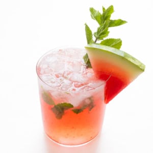 A refreshing watermelon mojito with ice, garnished with a slice of watermelon and a sprig of mint, against a white background.
