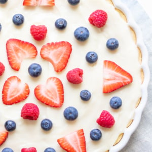 A fruit pizza with a creamy filling, topped with a pattern of fresh strawberries, raspberries, and blueberries.