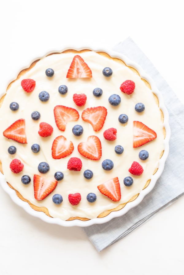 A fresh fruit pizza with cream and a decorative arrangement of strawberries, blueberries, and raspberries, finished with a glaze.