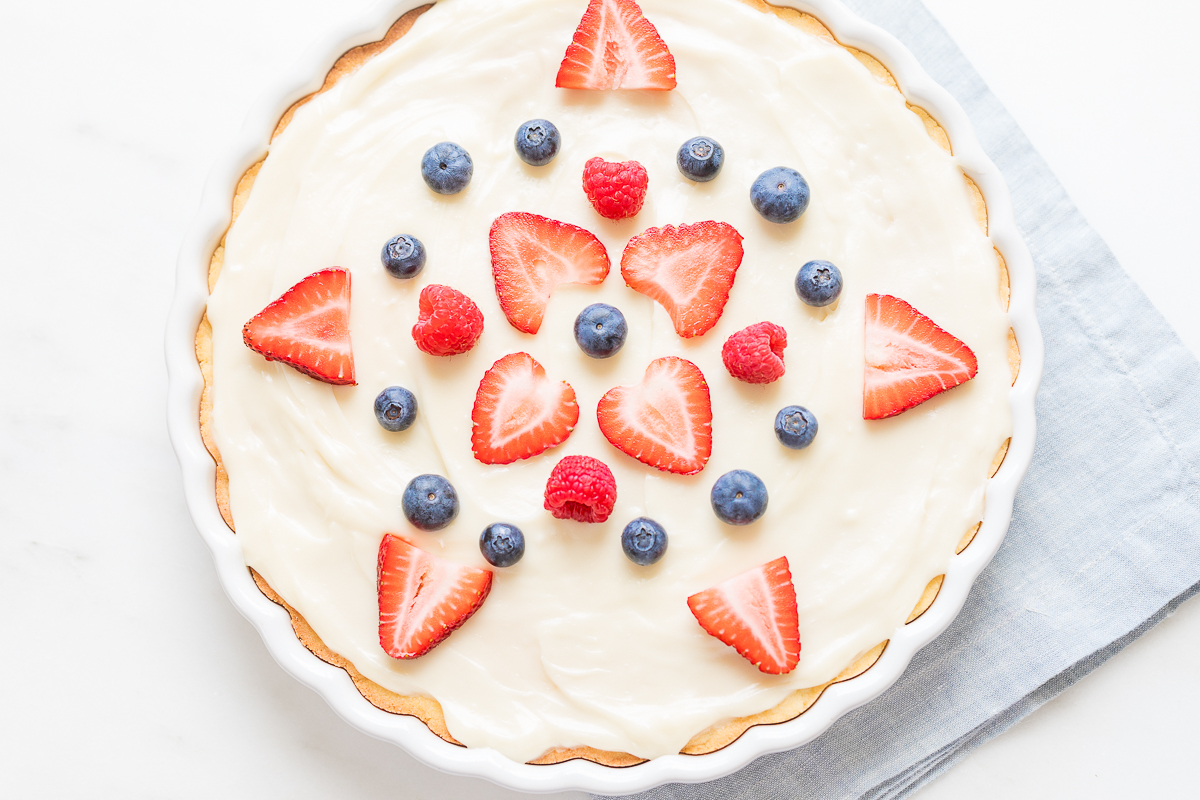 A freshly made fruit pizza crust topped with cream and garnished with strawberries and blueberries.