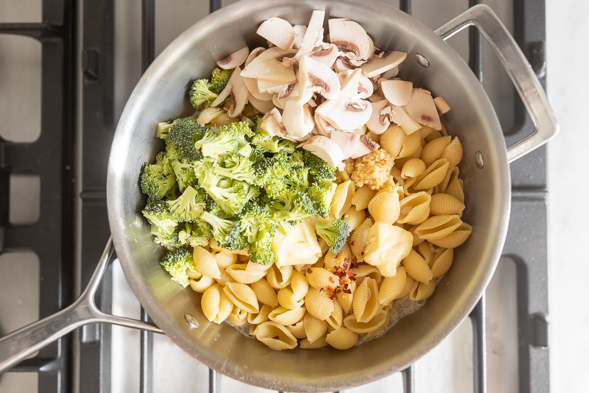 A stainless steel pan on a stove top with pasta shells and other ingredients to make pasta con broccoli.