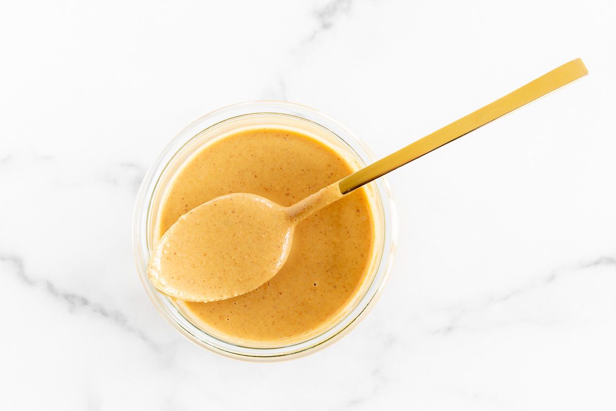 A glass jar full of homemade peanut butter on a marble surface, spoon resting across top