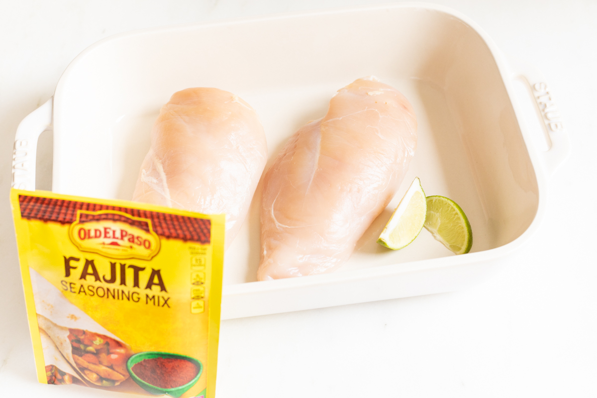 Chicken breasts in a white baking dish before baking. A pack of fajita seasoning is in the foreground.