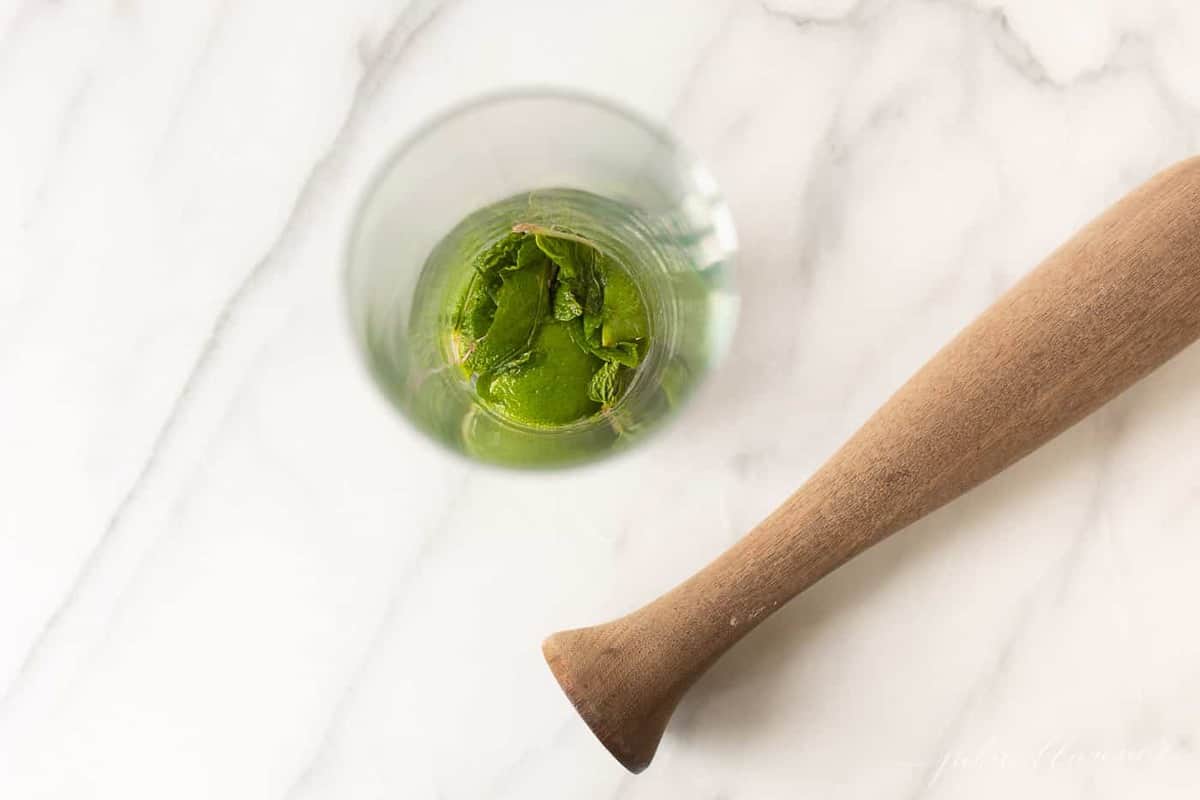 looking into a clear glass with mint on the bottom, next to a wooden muddler preparing to muddle the mint leaves for a mojito recipe.