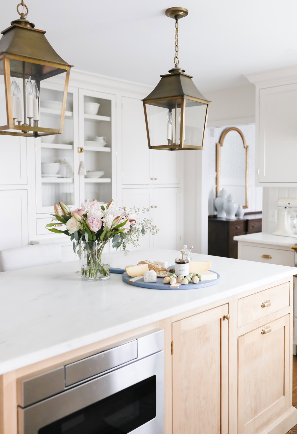 Elegant white kitchen with marble countertops, sophisticated lighting design, and a wooden island with a cheese platter and flowers.