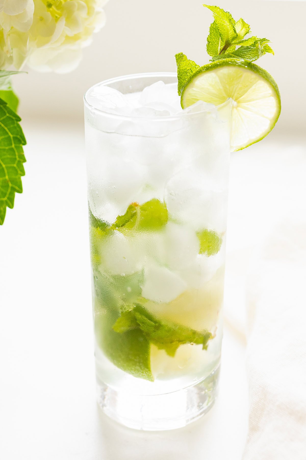 A mojito recipe in a clear glass with lime, mint, and ice on a white surface, garnished with a lime slice and mint leaf