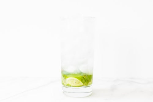 A tall glass of iced mojito with lime slices and mint leaves, set on a marble surface against a bright white background.