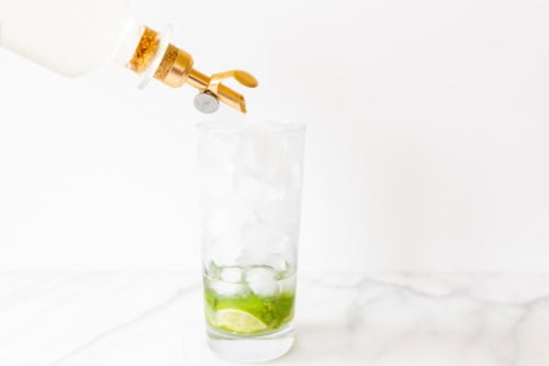 Pouring syrup from a bottle with a golden spigot into a tall glass filled with ice cubes and lime slices on a marble surface, crafting the perfect mojito.