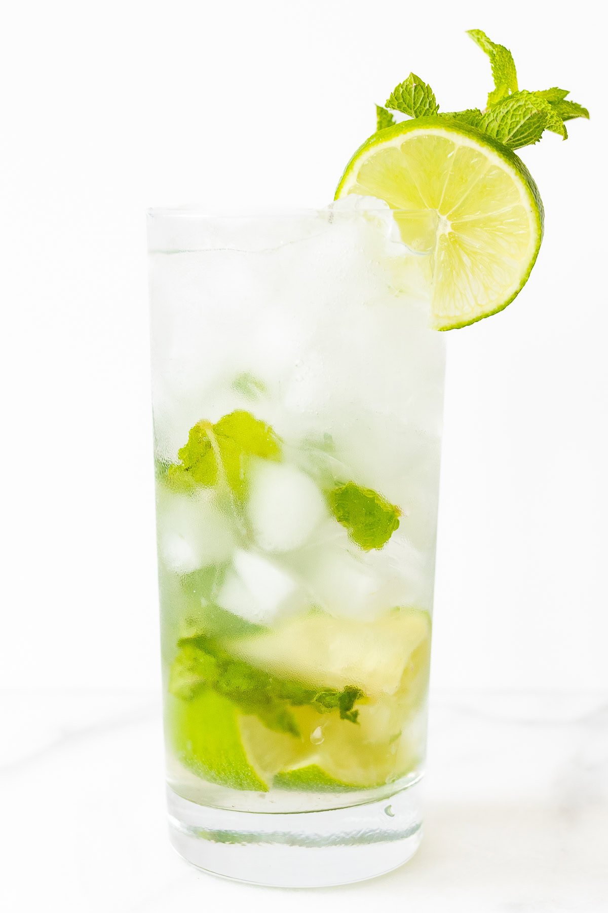 A glass of mojito with lime slices, ice cubes, and fresh mint leaves, garnished with a lime wedge and mint sprig on a white background.