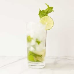 A classic mojito recipe in a clear glass on a marble surface, garnished with sprig of mint and a slice of lime.
