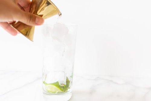 Pouring a mojito from a golden tin into a glass with ice cubes and lime slices on a marble countertop.