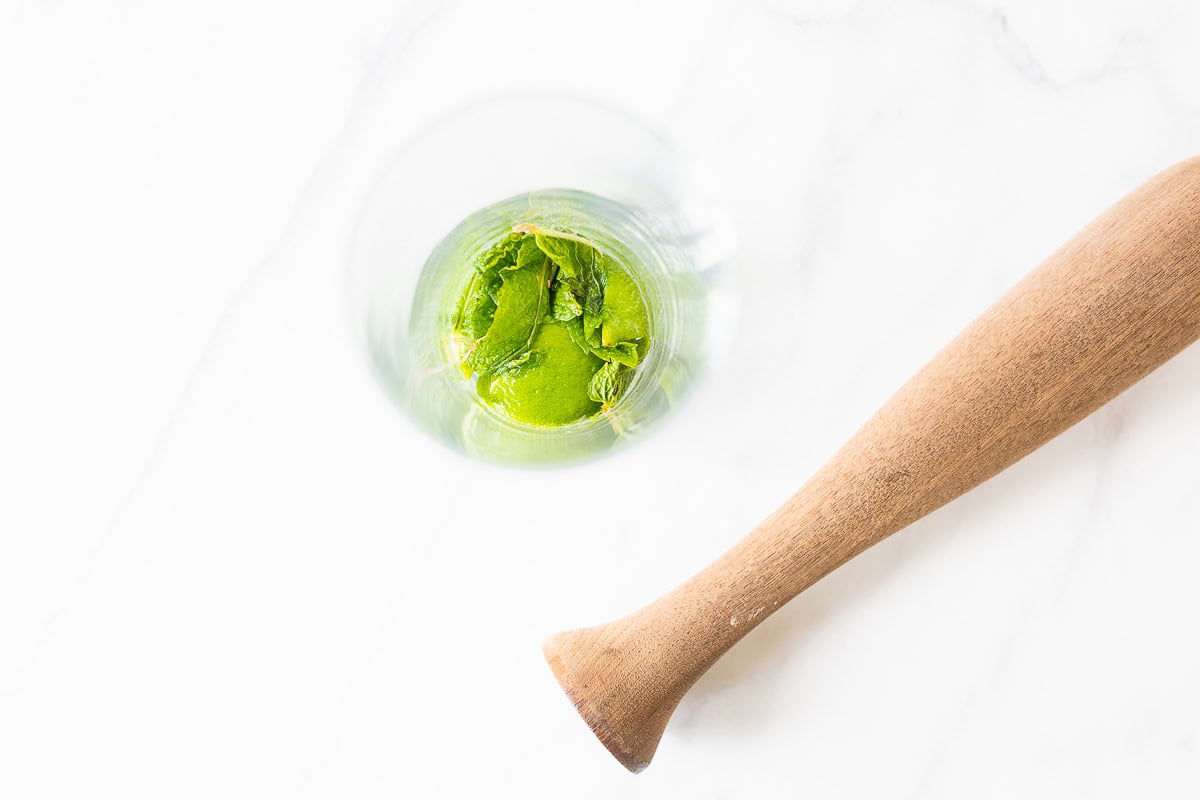 A clear glass with crushed mint leaves for a mojito recipe, with a wooden pestle resting beside it on a marble surface.