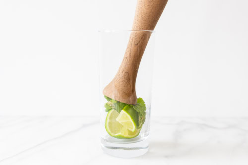 A wooden muddler pressing lime slices and mint leaves for a mojito recipe in a clear glass on a white marble background.