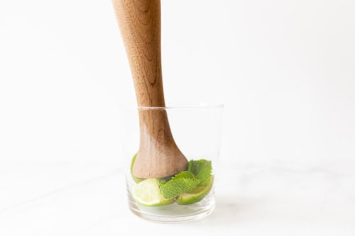 A wooden muddler presses lime slices and mint leaves at the bottom of a clear glass for a Skinny Mojito against a white background.