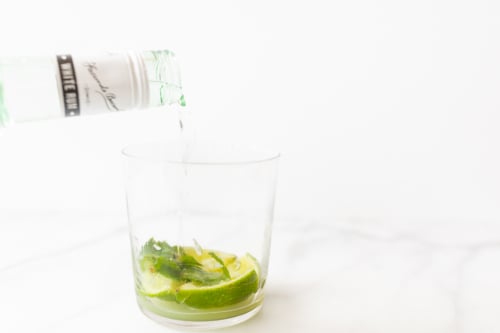 Rum being poured into a glass containing lime slices and mint leaves on a marble surface.