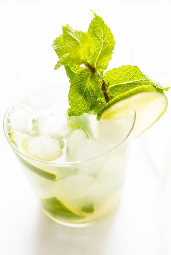 A clear glass filled with a skinny mojito cocktail, including ice cubes, fresh mint leaves, and a lime wedge on a bright white background.