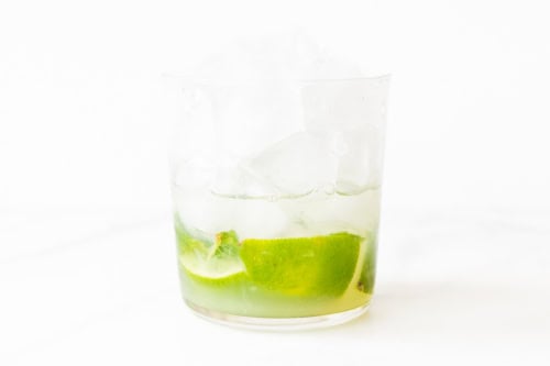 A clear glass containing ice cubes and a slice of lime, with a translucent liquid called Skinny Mojito, set against a white background.