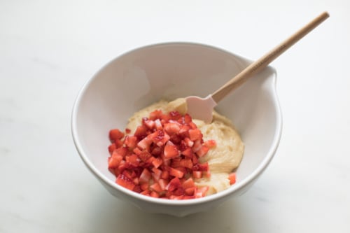 A bowl containing a blend of cream and diced strawberries with a wooden spoon on a marble surface.