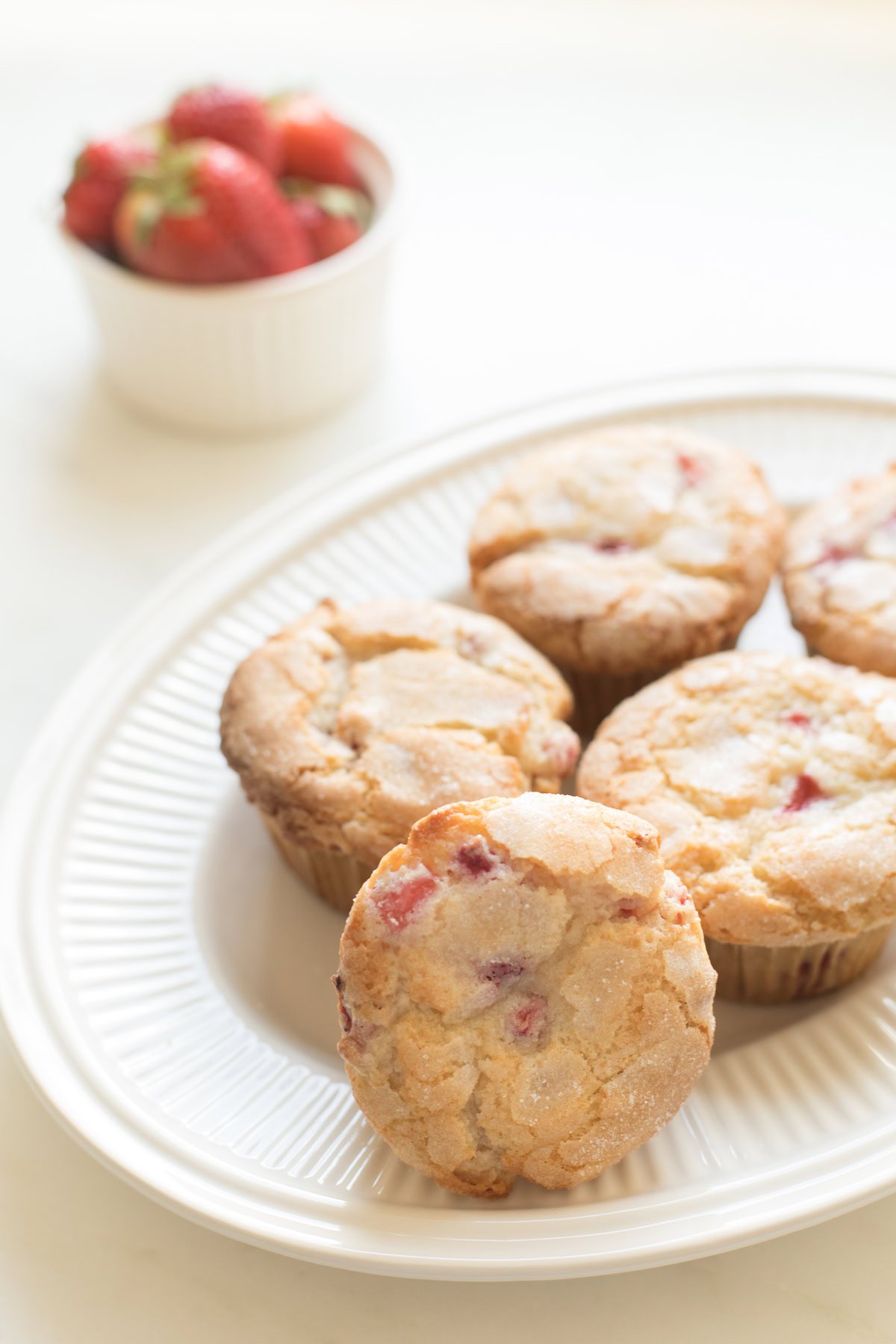 A plate with freshly baked strawberry muffins, one strawberry muffin prominently displayed in front, with a bowl of strawberries in the background.