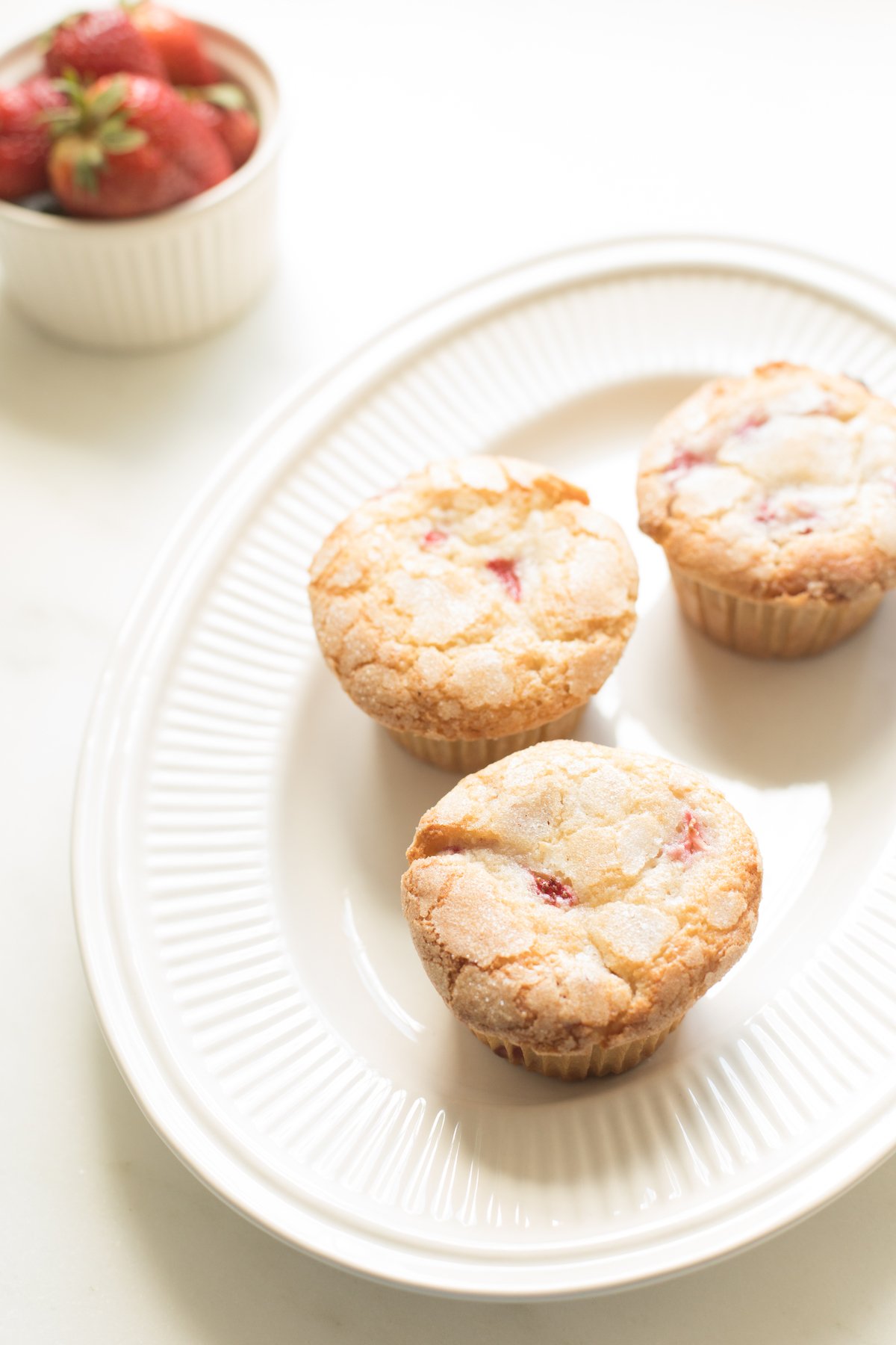 A plate of strawberry muffins with fresh strawberries in a cup on a white table.