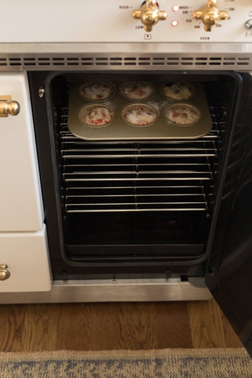 A modern oven with a batch of strawberry muffins baking on the middle rack, viewed from the front with the oven door open.