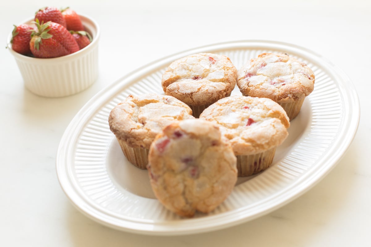 A plate of strawberry muffins with fresh strawberries in a cup on a white table.