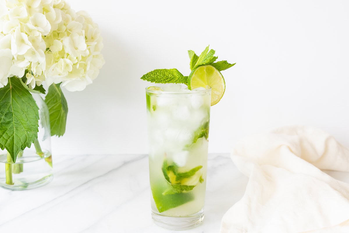 A refreshing vodka mojito cocktail with lime and mint leaves in a tall glass, accompanied by a hydrangea bouquet on a white marble surface.