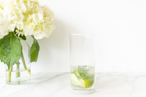 A glass of vodka mojito with lime slices on a white countertop, next to a vase of white hydrangeas.