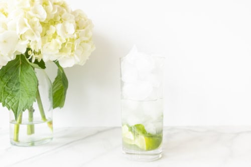 A glass of vodka mojito with ice and lime slices next to a vase with hydrangea flowers on a marble countertop.